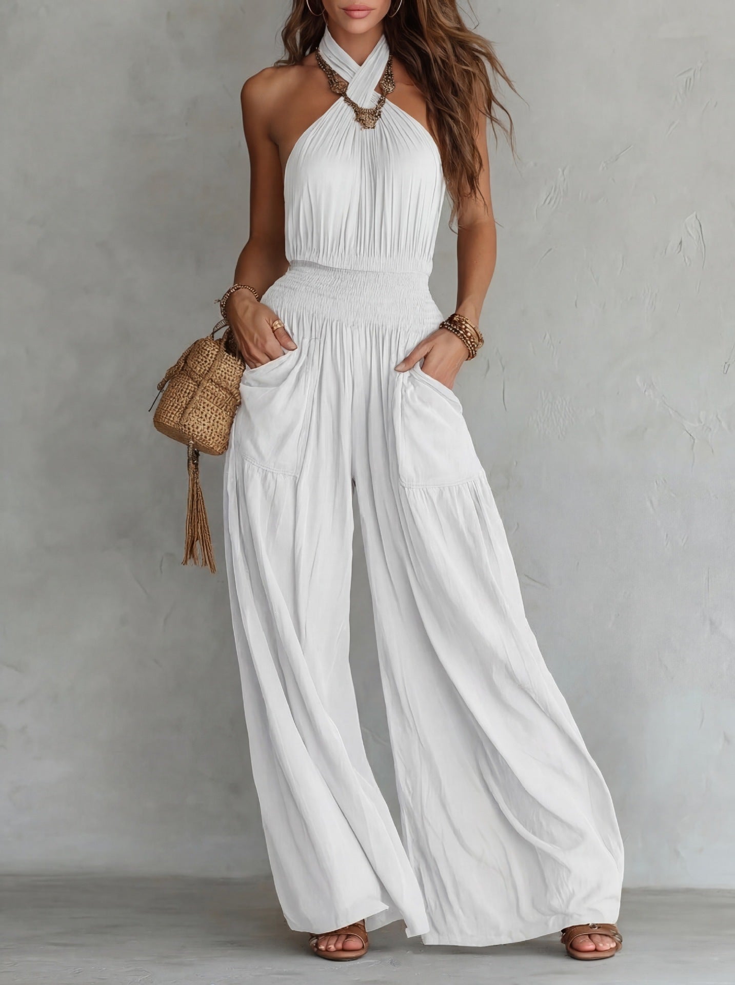 White bohemian sleeveless jumpsuit with halter neck, wide legs, drawstring waist, and front pockets. Model holds woven bag.